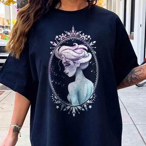 May include: A navy blue t-shirt featuring a detailed illustration of a woman with white hair and a blue-toned complexion, set within an oval frame. The frame is adorned with a decorative border of snowflakes and crystals.
