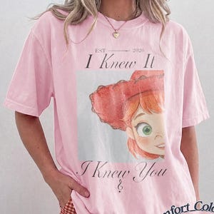 May include: A light pink short-sleeve t-shirt featuring a graphic print of a cartoon character with red hair and a red hat. The text reads "EST 2026 I Knew It I Knew You" in a serif font. The shirt has a crew neckline and a relaxed, loose fit.