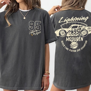 May include: Dark gray t-shirt featuring the text "95 Lightning McQueen" on the front and a graphic of a race car with the text "Lightning McQueen" and "Seven Time Piston Cup Champion" on the back. The shirt has a vintage, worn look.