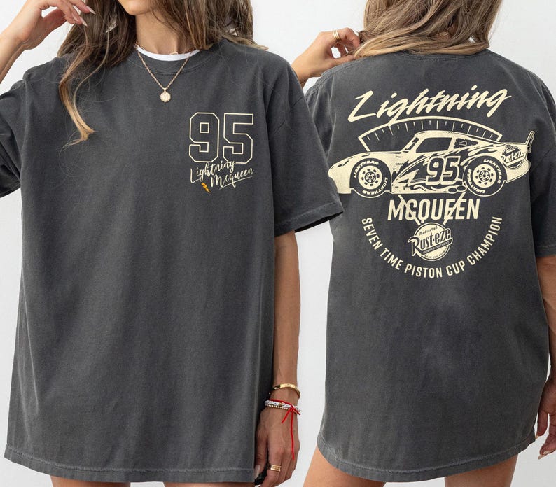 May include: Dark gray t-shirt featuring the text "95 Lightning McQueen" on the front and a graphic of a race car with the text "Lightning McQueen" and "Seven Time Piston Cup Champion" on the back. The shirt has a vintage, worn look.