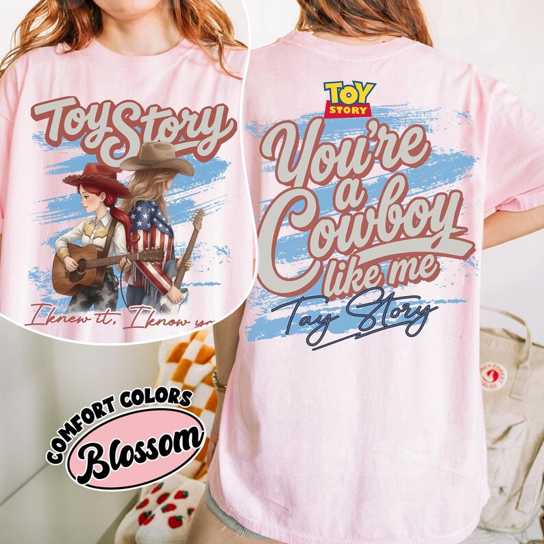 Może przedstawiać: Jasnor&oacute;żowy t-shirt z nadrukiem graficznym przedstawiającym dwie postacie z gitarami, napisem Toy Story oraz frazą You're a Cowboy like me. Projekt zawiera niebieskie akcenty w formie pociągnięć pędzla oraz małą grafikę z etykietą Comfort Colors Blossom.