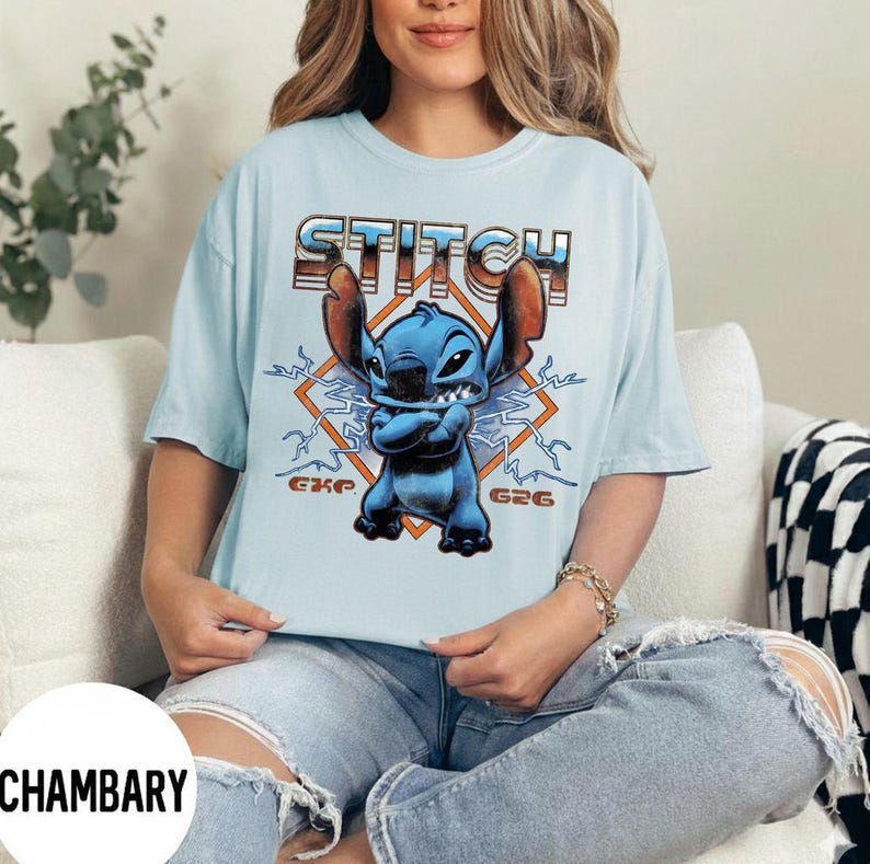 May include: Light blue t-shirt featuring a graphic of the character Stitch with the word "STITCH" in orange and brown lettering. The design includes lightning bolts and the text "G26" and "EXP". The shirt is a short-sleeved, casual top.