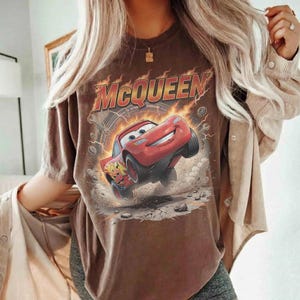 May include: Brown t-shirt featuring a graphic of Lightning McQueen from the movie Cars. The design includes the name "McQueen" in a fiery font above the red race car. The shirt has a vintage, distressed look.
