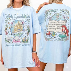 May include: Light blue t-shirts with a whimsical design. The front features a castle, musical notes, and text that reads "Wish I could be Part of That World." The back showcases a mermaid, sheet music, and aquatic elements.