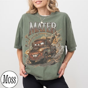 K&ouml;nnte beinhalten: Olivgr&uuml;nes T-Shirt mit einem Aufdruck von Mater aus dem Film Cars. Das Design umfasst das Wort "MATER", Blitze und Werkzeuge. Der Zeichentrick-Abschleppwagen ist braun mit einem l&auml;chelnden Gesicht, vor einem verwitterten Hintergrund.