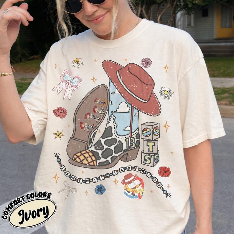 May include: An ivory t-shirt featuring a graphic print of a brown cowboy hat, a cow-print cowboy boot with a blue sky pattern, alphabet blocks, and a beaded string with the text I KNEW IT I KNEW YOU, surrounded by small floral and star icons.