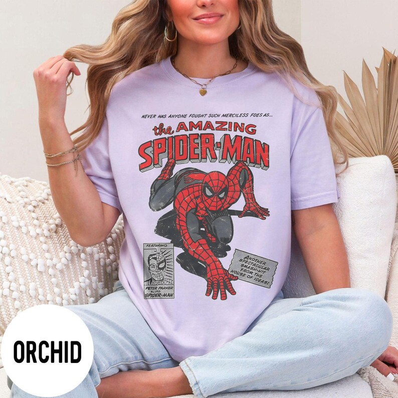 May include: A light purple t-shirt featuring a vintage comic book graphic of Spider-Man. The design includes the text "the AMAZING SPIDER-MAN" in red and blue, with additional text and a comic book style illustration of Spider-Man.