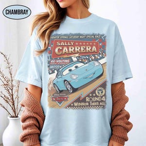 May include: Light blue t-shirt featuring a graphic of Sally Carrera from the Disney Pixar Cars movie. The design includes text that reads "Radiator Springs Saturday Night Special Race" and "Winner Takes All."