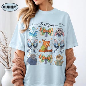 May include: A light blue t-shirt featuring the text Zootopia and a graphic print of various characters from the movie, including a fox, a rabbit, a buffalo, and a beaver, interspersed with several colourful patterned bows and small stars.