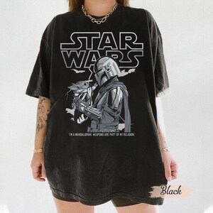 Może przedstawiać: Czarny t-shirt Star Wars z grafiką Mandalorianina i Grogu. Logo Star Wars znajduje się nad ilustracją. Tekst "I'M A MANDALORIAN. WEAPONS ARE PART OF MY RELIGION." znajduje się poniżej.