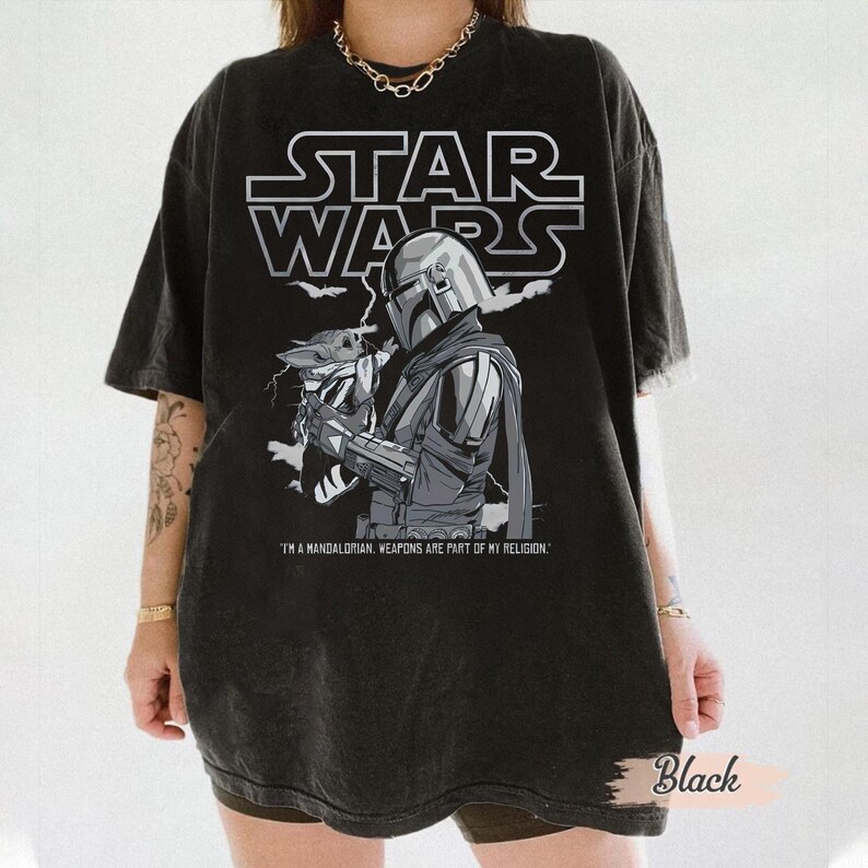 Może przedstawiać: Czarny t-shirt Star Wars z grafiką Mandalorianina i Grogu. Logo Star Wars znajduje się nad ilustracją. Tekst "I'M A MANDALORIAN. WEAPONS ARE PART OF MY RELIGION." znajduje się poniżej.