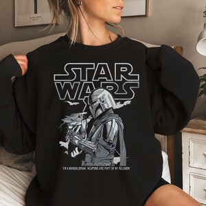 Może przedstawiać: Czarna bluza z nadrukiem Star Wars. Grafika zawiera logo Star Wars, postać Mandaloriana i Grogu. Tekst brzmi: "JESTEM MANDALORIANINEM. BROŃ JEST CZĘŚCIĄ MOJEJ RELIGII."