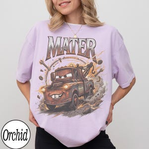 K&ouml;nnte beinhalten: Helllila T-Shirt mit einem Cartoon-Bild von Hook aus dem Film Cars. Das Wort "MATER" ist in einer verwitterten Schriftart &uuml;ber dem Bild aufgedruckt. Das Bild enth&auml;lt Schraubenschl&uuml;ssel und ein kreisf&ouml;rmiges Design.