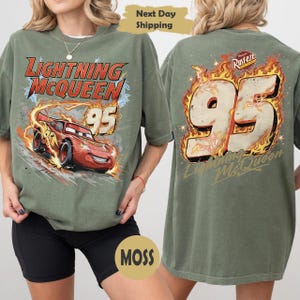 May include: Olive green t-shirt featuring Lightning McQueen from the Cars film. The front graphic shows the car with the number 95 and the text "Lightning McQueen". The back has a large "95" in flames.