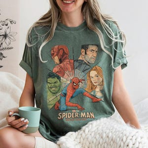 May include: Green t-shirt featuring a graphic of Spider-Man and other Marvel characters. The design includes the text "Spider-Man: Brave New Day" in a vintage style. The shirt is a muted green color.