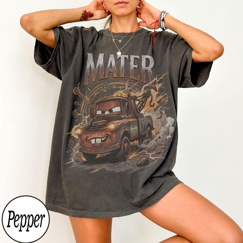 K&ouml;nnte beinhalten: Dunkelgraues T-Shirt mit dem Wort "MATER" &uuml;ber einem Cartoon-Bild von Abschleppwagen Hook aus dem Film Cars. Die Grafik ist in Braun-, Orange- und Graut&ouml;nen gehalten. Das Shirt hat kurze &Auml;rmel und eine lockere Passform.