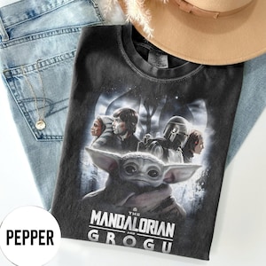 Op de afbeelding: Een zwart t-shirt met personages uit de serie "The Mandalorian", waaronder Grogu. Het shirt heeft de tekst "The Mandalorian and Grogu" in het wit. Het shirt ligt op een blauwe spijkerbroek, een ketting en een beige hoed.