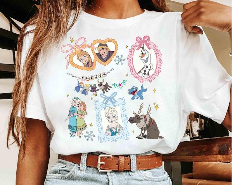 以下が含まれることがあります： 映画「アナと雪の女王」のキャラクターが描かれた、カラフルなイラストが特徴の白いTシャツ。デザインには、アナ、エルサ、オラフ、クリストフ、スヴェンの画像が含まれており、装飾的な枠で囲まれています。「FROZEN」の文字も表示されています。