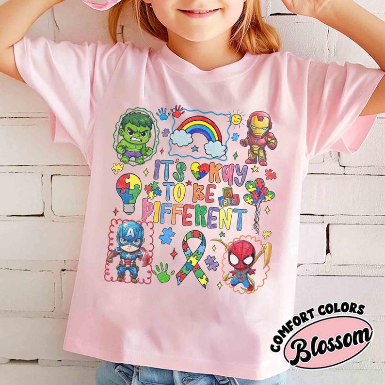 K&ouml;nnte beinhalten: Ein hellrosa T-Shirt mit Cartoon-Illustrationen von Superhelden und einem Regenbogen. Das Shirt tr&auml;gt den Text "IT'S OKAY TO BE DIFFERENT" und ein Band mit Puzzleteilen.