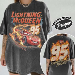 May include: Dark grey t-shirt featuring Lightning McQueen from the Cars film. The front graphic shows McQueen with the number 95 and flames. The back of the shirt also has the number 95 and the text "Lightning McQueen". The shirt is labelled "Comfort Colors Pepper".