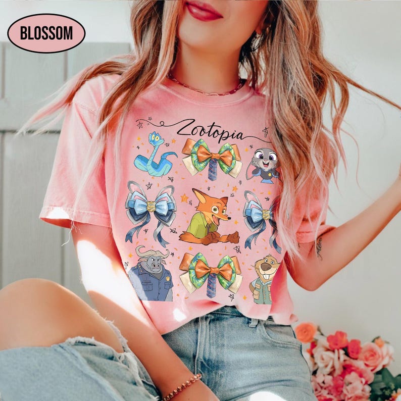 May include: A coral pink short-sleeve t-shirt featuring a Zootopia graphic print. The design includes characters like Nick Wilde, Judy Hopps, and Chief Bogo, interspersed with decorative bows and stars. The text Zootopia is printed in black script.