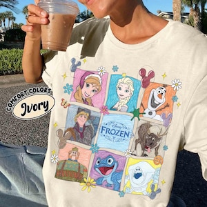 May include: Ivory t-shirt with a colourful graphic of characters from Disney's Frozen. The design includes images of Anna, Elsa, Olaf, Kristoff, Sven, and other characters. The shirt also has the words "Disney Frozen" and "Comfort Colors Ivory".