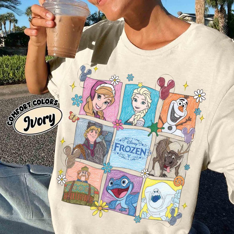 May include: Ivory t-shirt with a colourful graphic of characters from Disney's Frozen. The design includes images of Anna, Elsa, Olaf, Kristoff, Sven, and other characters. The shirt also has the words "Disney Frozen" and "Comfort Colors Ivory".