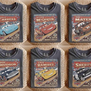 May include: Six gray t-shirts featuring characters from the movie Cars. Each shirt displays a different character: Sally Carrera, Lightning McQueen, Tow Mater, Doc Hudson, Cruz Ramirez, and Sheriff. The shirts have a vintage race-themed design.
