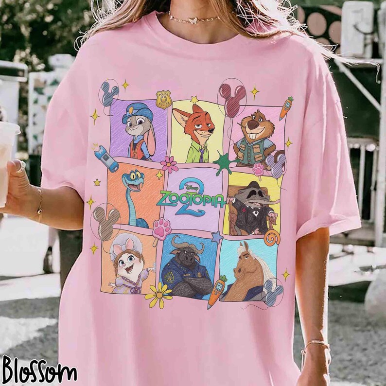 Puede incluir: Camiseta rosa claro con un dise&ntilde;o gr&aacute;fico colorido de personajes de la pel&iacute;cula "Zootopia 2". El dise&ntilde;o incluye a Judy Hopps, Nick Wilde y otros personajes en un patr&oacute;n de cuadr&iacute;cula. La palabra "Blossom" est&aacute; impresa abajo a la izquierda.