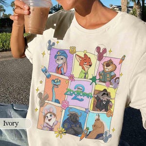 Puede incluir: Camiseta de color marfil con un gr&aacute;fico colorido de personajes de la pel&iacute;cula de Disney Zootopia. El dise&ntilde;o incluye a Judy Hopps, Nick Wilde y otros personajes en un patr&oacute;n de cuadr&iacute;cula. La camiseta est&aacute; hecha de un material suave.