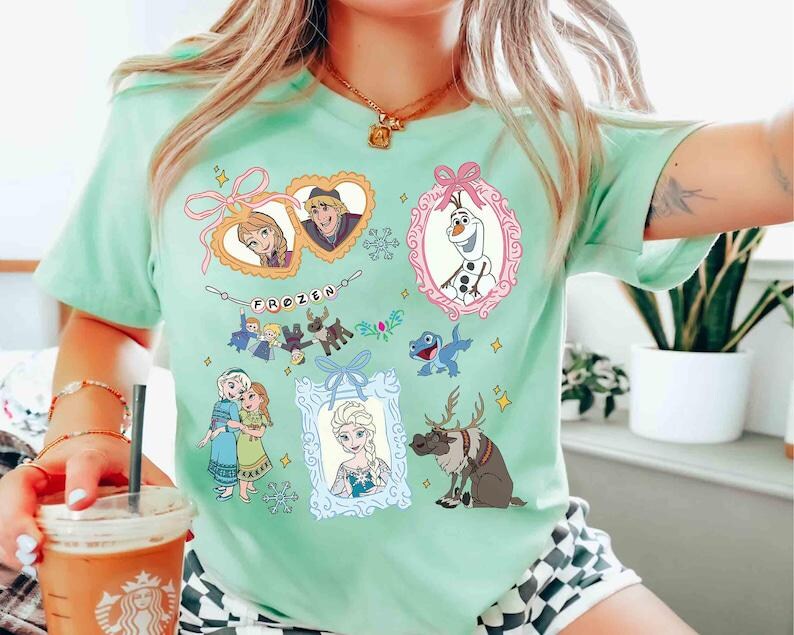 以下が含まれることがあります： 映画「アナと雪の女王」のキャラクターが描かれた、薄い緑色のTシャツ。アナ、エルサ、クリストフ、オラフ、スヴェンの額縁に入った肖像画と、上部に「FROZEN」の文字がプリントされています。