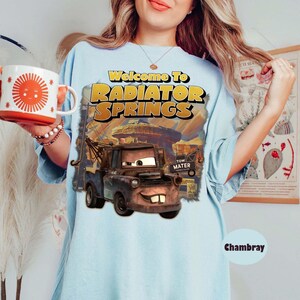 May include: A light blue t-shirt featuring a graphic print of the character Tow Mater from Radiator Springs. The text reads Welcome To Radiator Springs and Tow Mater. A white mug with a red sun graphic is held in the foreground of the image.