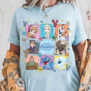 May include: Pale blue t-shirt with a colourful, illustrated grid of characters from Disney's Frozen. The grid includes Anna, Elsa, Olaf, Kristoff, Sven, and other characters, with the word "Frozen" in the centre.