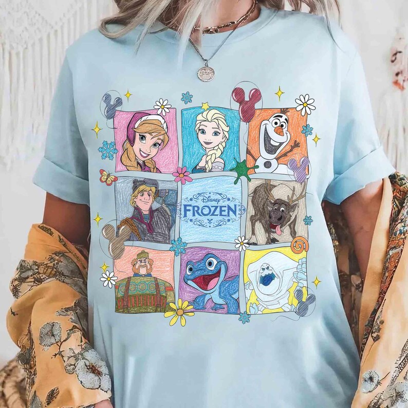 May include: Pale blue t-shirt with a colourful, illustrated grid of characters from Disney's Frozen. The grid includes Anna, Elsa, Olaf, Kristoff, Sven, and other characters, with the word "Frozen" in the centre.