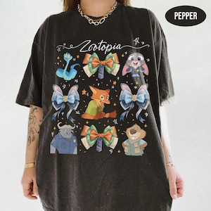 May include: A black short-sleeve t-shirt featuring a graphic print with the text Zootopia, various character illustrations including a fox, rabbit, and buffalo, and several decorative bows interspersed with small star shapes. The shirt colour is Pepper.