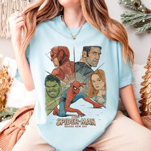 May include: Light blue t-shirt featuring a graphic of Spider-Man and other Marvel characters. The design includes Daredevil, Hulk, and The Punisher. The text "SPIDER-MAN BRAND NEW DAY" is printed below the characters.