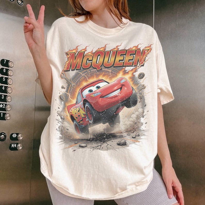 May include: Cream-colored t-shirt featuring a graphic of Lightning McQueen from the Cars movie. The design includes the word "McQueen" in a fiery font above the red race car. The shirt has short sleeves and a relaxed fit.