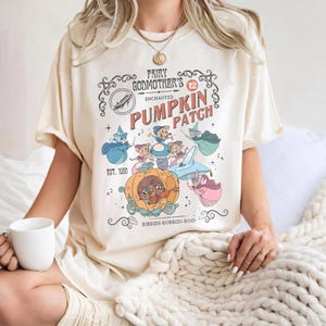 May include: Cream-colored t-shirt featuring a whimsical graphic of a pumpkin carriage with fairy godmothers and the text "Fairy Godmother's Enchanted Pumpkin Patch." The design includes the text "Est. 1950" and "Bibbidi-Bobbidi-Boo!"