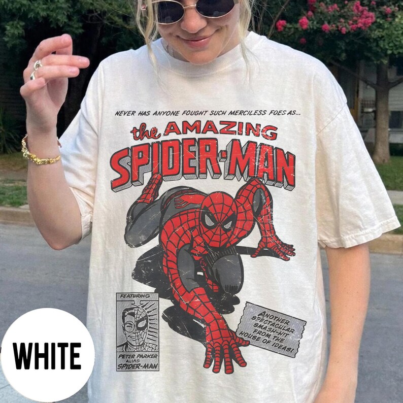 May include: White t-shirt featuring a graphic of Spider-Man in red and black, with the text "the AMAZING SPIDER-MAN". Additional text includes "NEVER HAS ANYONE FOUGHT SUCH MERCILESS FOES AS..." and "ANOTHER SPECTACULAR SMASH-HIT FROM THE HOUSE OF IDEAS!"