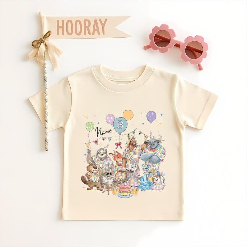 May include: A cream-colored t-shirt featuring a colorful illustration of cartoon animals celebrating a birthday. The number 3 is on a balloon. Pink flower-shaped sunglasses and a banner that says "HOORAY" are also in the image.
