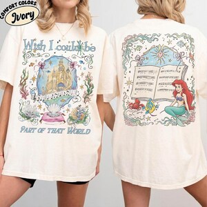 May include: Ivory-colored t-shirt featuring a colorful illustration. The front design includes a castle, musical notes, and the text "Wish I could be Part of That World." The back shows a mermaid, sheet music, and sea creatures.