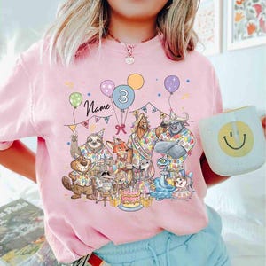 May include: Pink t-shirt featuring a colorful illustration of cartoon animals celebrating a birthday. The design includes balloons, a cake, and the number 3. The word "Name" is written above the animals.