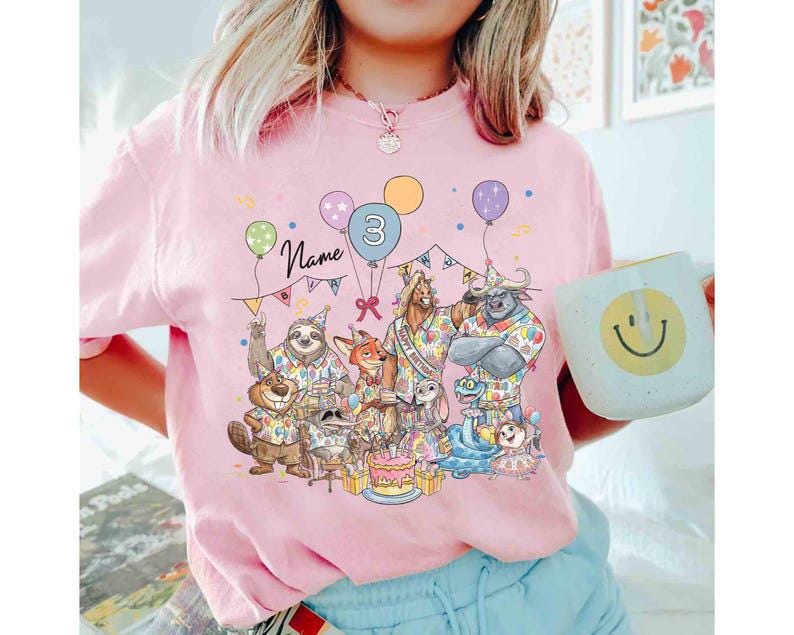 May include: Pink t-shirt featuring a colorful illustration of cartoon animals celebrating a birthday. The design includes balloons, a cake, and the number 3. The word "Name" is written above the animals.