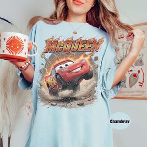 May include: Light blue t-shirt featuring a graphic of Lightning McQueen from the movie Cars. The design includes the name "McQueen" in fiery lettering above the red race car. The shirt also has the number 95 on the car.
