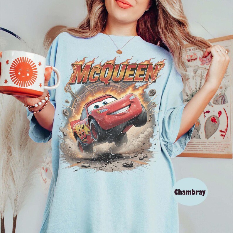 May include: Light blue t-shirt featuring a graphic of Lightning McQueen from the movie Cars. The design includes the name "McQueen" in fiery lettering above the red race car. The shirt also has the number 95 on the car.