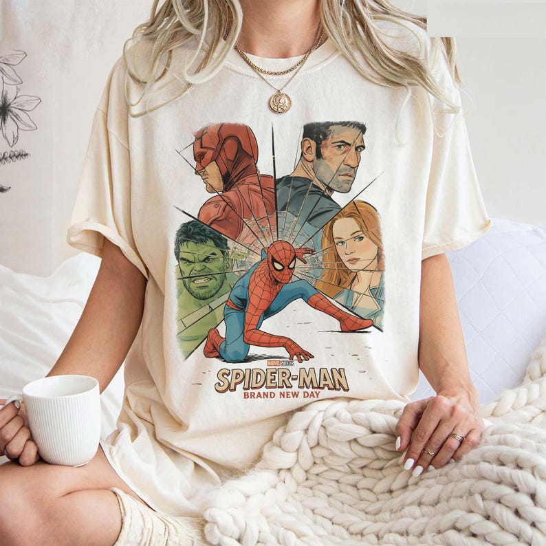 May include: Cream-colored t-shirt featuring a graphic of Spider-Man and other Marvel characters. The design includes the text "SPIDER-MAN BRAND NEW DAY" in a vintage style. The shirt is made of a soft material.