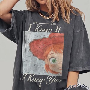 May include: A charcoal gray short-sleeve t-shirt featuring a graphic print of a red-haired character wearing a red hat. The text reads "EST 2026 I Knew It I Knew You" with a small treble clef symbol. The shirt has a distressed, vintage-style wash.