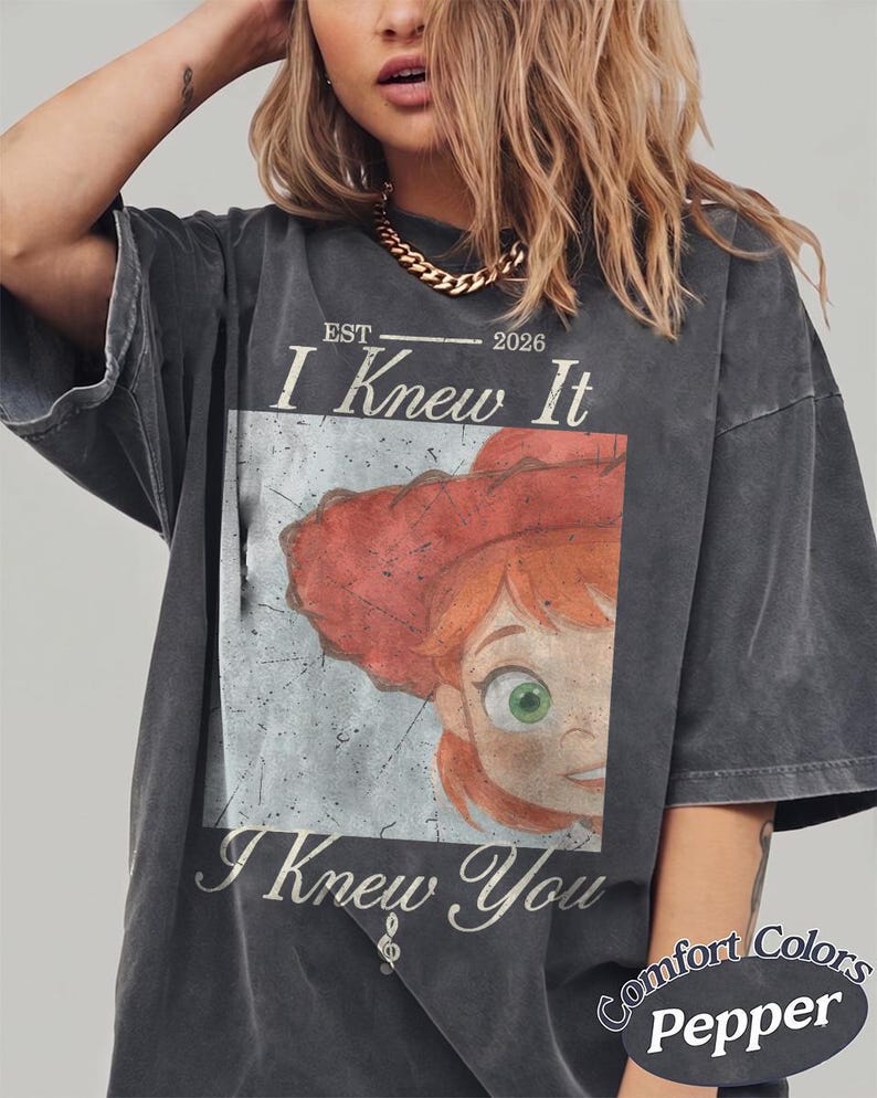 May include: A charcoal gray short-sleeve t-shirt featuring a graphic print of a red-haired character wearing a red hat. The text reads "EST 2026 I Knew It I Knew You" with a small treble clef symbol. The shirt has a distressed, vintage-style wash.