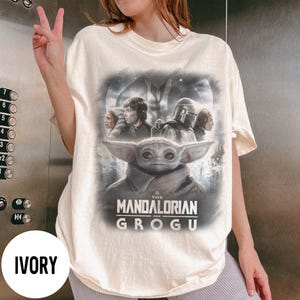Op de afbeelding: Ivoor kleurig T-shirt met een afbeelding uit de serie "The Mandalorian". Het ontwerp bevat Grogu, de Mandalorian en andere personages. De tekst "The Mandalorian and Grogu" staat onder de personages.