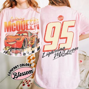 May include: A light pink t-shirt featuring Lightning McQueen from the Cars movie. The front graphic shows the car with the text "Lightning McQueen" and "95". The back of the shirt displays a large "95" and "Lightning McQueen".